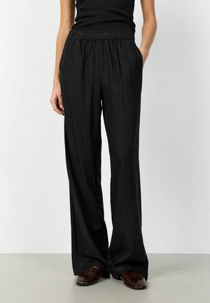 Femeie purtând pantaloni negri largi cu talie elastică și pantofi maro închis, stând cu o mână în buzunar, pe fundal alb.