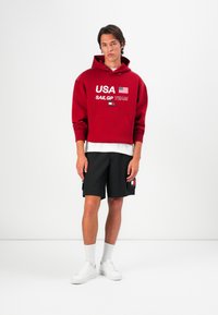 Rød hættetrøje med "USA" og flagdesign, parret med sorte shorts med sidelommer og logo-patch, afsluttet med hvide sneakers.