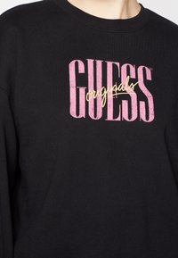 Svart sweatshirt med en rosa och gul "GUESS" logotyp. Tyget är slätt med en klassisk rundad halsringning och en avslappnad passform.