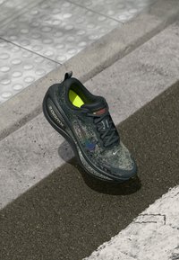 Dunkelgrauer Nike ZoomX-Laufschuh mit schwarzen Schnürsenkeln und leuchtend gelber Innenseite auf strukturiertem urbanem Pflaster.