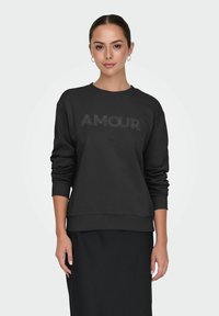 Joven mujer vestida con sudadera negra que tiene el texto "AMOUR" y falda negra, de pie contra un fondo liso claro, mirando hacia adelante.