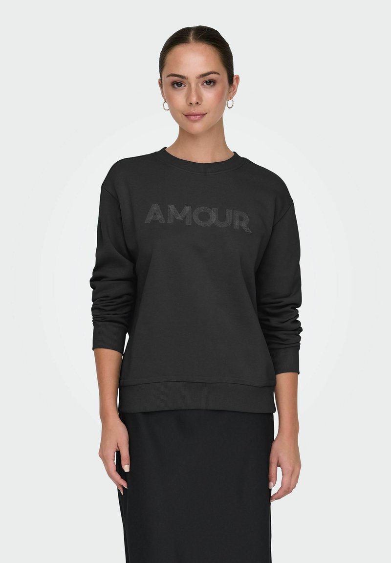 Joven mujer vestida con sudadera negra que tiene el texto "AMOUR" y falda negra, de pie contra un fondo liso claro, mirando hacia adelante.