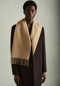 Beige villane sall, millel on frantsi lõpuotsad, on visatud tumeda pruuni jope peale. Tekstuur tundub pehme ja soe, lihtsalt ning elegantse disainiga.