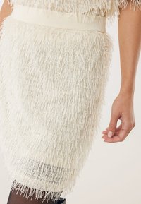 Sukienka z frędzlami w odcieniu białym, z teksturowanej tkaniny z długimi, luźnymi pasmami i gładkim pasem w talii. Długość dołem lekko powyżej kolana.