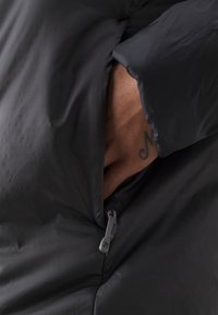 Black padded jacket with a close-up of a hand in a side pocket. Features a smooth texture, zipper closure, and a gray drawstring.