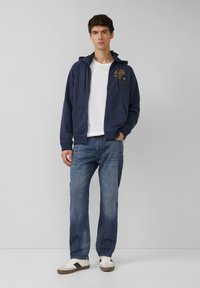 Granatowa bluza z zamkiem i logo z przodu, zestawiona z jasnoniebieskimi jeansami o prostych nogawkach oraz białymi tenisówkami z szarymi i czarnymi akcentami.