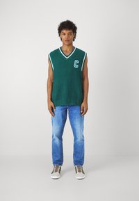 Un pulover verde cu dungi, fără mâneci, cu tiv alb și litera „C” pe piept, asortat cu blugi blue deschis și adidași gri.