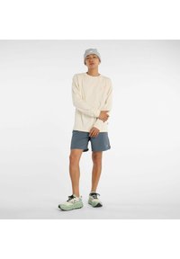 Cremefarbiges Langarmshirt mit pinkem Text, kombiniert mit blauen Sportshorts. Die Schuhe haben ein mehrfarbiges Design mit Schnürsenkeln.