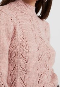 Maglione lavorato a maglia chunky color rosa con colletto alto, caratterizzato da un motivo a zig zag testurizzato e dettagli a coste sul collo e sui polsini.