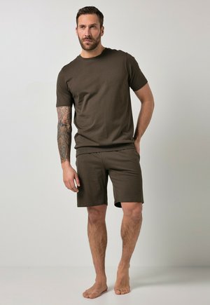 Hombre con el brazo izquierdo tatuado que lleva una camiseta marrón oscura y pantalones cortos a juego, de pie descalzo sobre un fondo claro y liso.