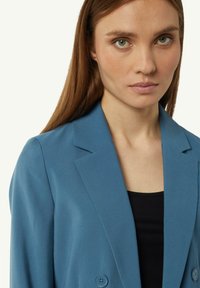 Niebieska marynarka dopasowana z kołnierzem z klapami, podwójnymi guzikami i prążkowaną teksturą. Nosi się ją na czarnym topie, podkreślającym dopasowaną sylwetkę.