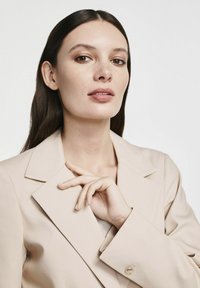 Beżowa marynarka o dopasowanym kroju z klapami, gładką teksturą i ozdobnym guzikiem na rękawie. Model delikatnie dotyka marynarki.