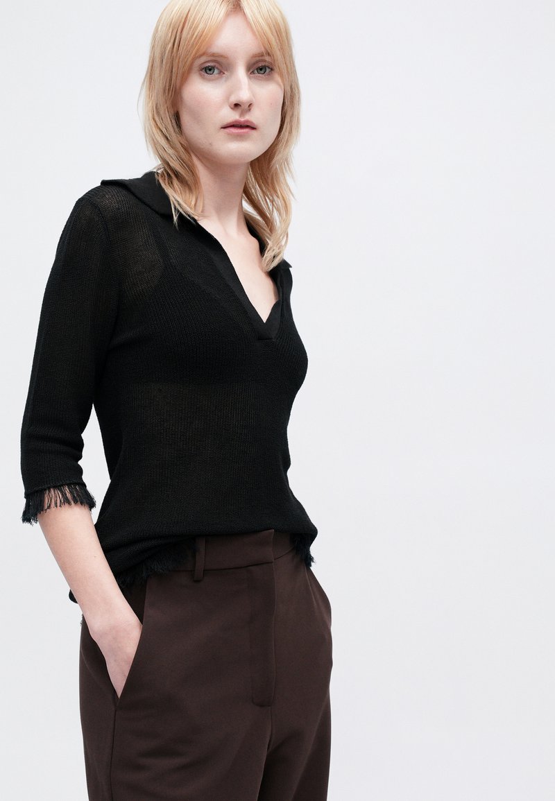 Top in maglia nera con scollatura a V e colletto, dotato di maniche a tre quarti con bordi frangiati. Abbinato a pantaloni tailleur marroni.