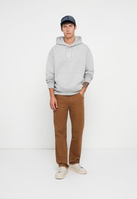 Sudadera gris con bolsillo frontal y logo bordado; pantalones rectos marrones; zapatillas blancas; gorra azul marino con un patrón texturizado.