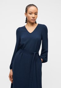 Νέα γυναίκα με πλεξούδες, φορώντας μακρυμάνικο φόρεμα σε ναυτικό μπλε χρώμα με V λαιμόκοψη και υφασμάτινη ζώνη, στέκεται μπροστά σε απλό φόντο.