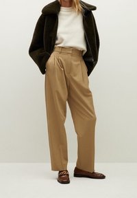 Beige plisserede bukser parret med en hvid sweater og en mørkegrøn fleecejakke, fuldendt med brune loafers med en guld kædeaccent.