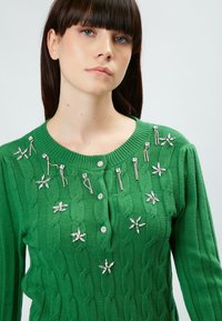 Grüner gestrickter Cardigan mit Kabelmuster, verziert mit silbernen floralen und nadelähnlichen Akzenten sowie einem runden Ausschnitt mit Knopfverschluss.