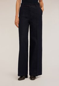 Široké tmavé džíny s předními kapsami, viditelným šitím a hnědým pásem, spárované s tmavými podpatky.