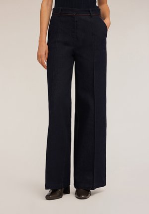 Pantaloni in denim scuro a gamba larga con tasche frontali, cuciture a vista, e una cintura marrone in evidenza, abbinati a scarpe con tacco scuro.