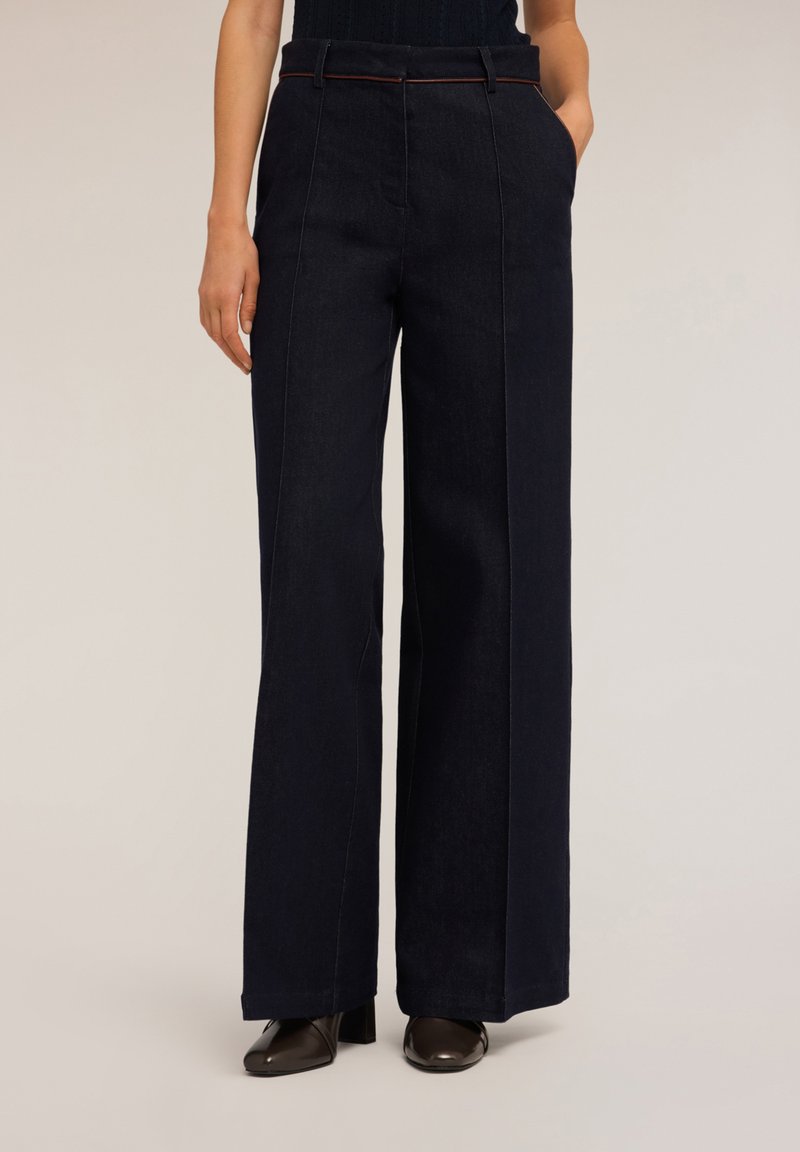 Široké tmavé džíny s předními kapsami, viditelným šitím a hnědým pásem, spárované s tmavými podpatky.