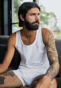 Hvid tanktop og hvide underbukser båret af en mand med langt hår og skæg. Bemærkelsesværdige tatoveringer på hans arme og en blød grå sofa i baggrunden.
