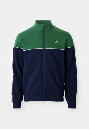 Full-zip jakke med grønn øvre del av bryst og skuldre, marineblå underdel og ermer, hvite kanter og liten krokodillelogo på brystet.