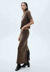 Haut court marron avec un col rond, associé à une longue jupe marron avec une fente sur le côté. Le mannequin porte des bottines noires à la cheville. Fond minimaliste.