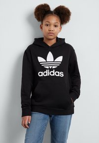 Mustmustelne kapuutsiga sviiter, millel on suur valge Adidas logo ja kolm riba disain rinnal. Ees on kenguru tasku, taustal hele taust.