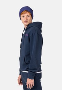 Felpa blu navy con tasca a marsupio, cordino regolabile e logo viola; abbinata a pantaloni coordinati e un berretto a coste viola.