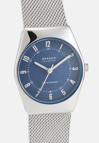 Zegarek na srebrnym siateczkowym pasku z heksagonalną tarczą, niebieskim cyferblatem, białymi znaczkami numerycznymi oraz funkcją zasilania słonecznego. Nazwa marki znajduje się na środku.