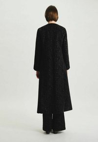Long manteau noir avec un motif texturé en dentelle. Il a des manches longues et une silhouette droite, associé à un pantalon noir évasé.