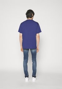 Camiseta de manga corta y cuello redondo en un azul oscuro sólido, combinada con unos jeans ajustados de mezclilla y zapatillas blancas, vista desde atrás.