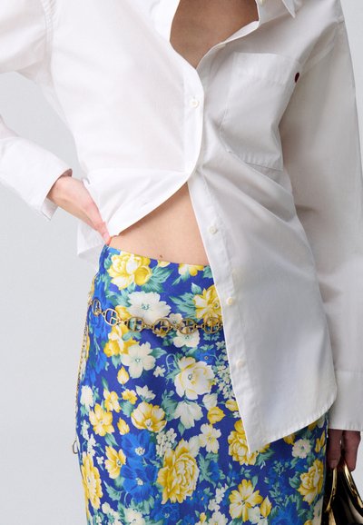 Chemise en coton blanche avec des boutons visibles, associée à une jupe florale bleue avec des accents jaunes et une ceinture en chaîne dorée.