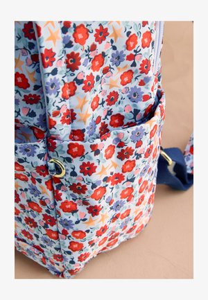 Tasca di uno zaino in tessuto con motivo floreale con fiori rossi e viola, stelle beige e una tracolla blu su uno sfondo beige.