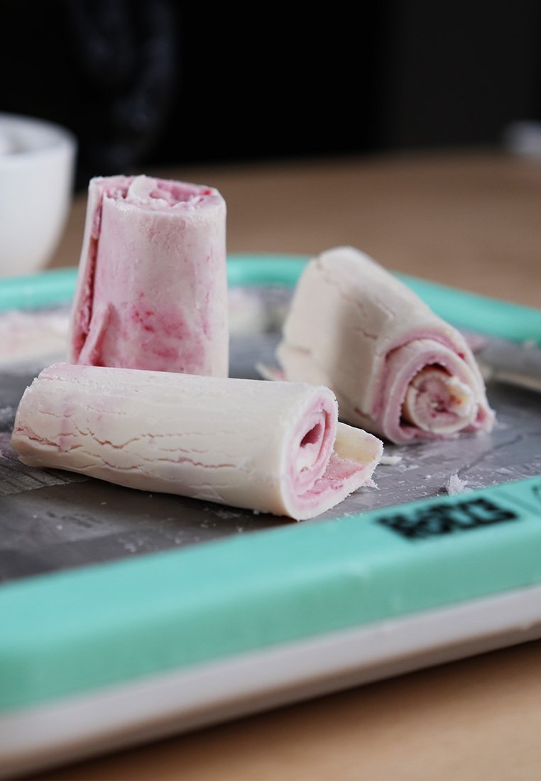 Rollen von gefrorenem Dessert mit rosa-weißen Wirbeln, auf einem Metalltablett mit türkisem Rand platziert, das eine glatte Textur und abgerundete Form zeigt.