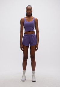 Γυναίκα στέκεται προς τα εμπρός φορώντας μωβ αθλητική φανέλα χωρίς μανίκια, ταιριαστά σορτς με λογότυπο Under Armour, άσπρες κάλτσες και άσπρα αθλητικά παπούτσια.