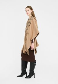 Poncho beige con colletto sciallato a motivo, indossato sopra un vestito aderente marrone, abbinato a stivali neri con tacco alti fino al ginocchio. Texture liscia, design casual.