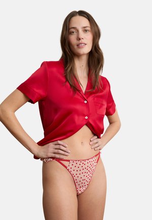 Femme portant un chemisier rouge en satin à manches courtes et un slip beige transparent avec des motifs de cœurs rouges, debout les mains sur les hanches devant un fond blanc.