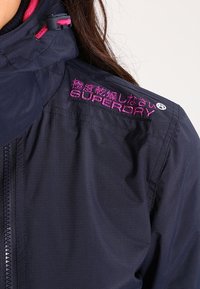 Marineblå vindbreakerjakke med tekstureret stof, med pink broderet "SUPERDRY" logo på skulderen og justerbar kravehardware.