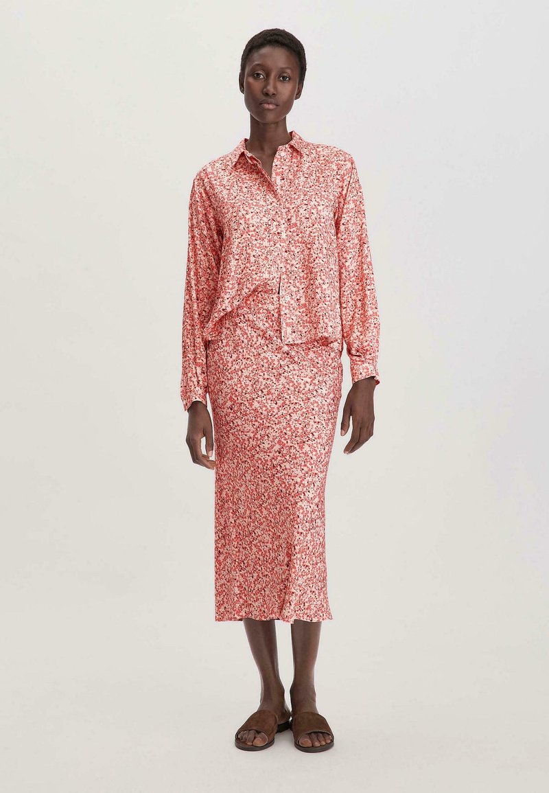 Bluse aus Satin mit floralem Muster in Rot und Weiß, mit durchgehender Knopfleiste, kombiniert mit einem passenden knielangen Rock. Das Model trägt braune Sandalen.