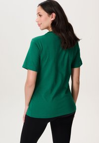 Vrouw met donker haar die zijwaarts kijkt, gekleed in een groene T-shirt met korte mouwen en zwarte broeken, tegen een effen achtergrond.