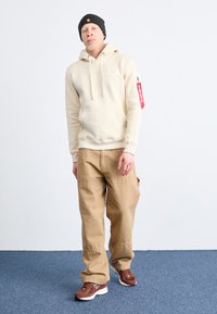 Sweatshirt à capuche beige avec poche avant, étiquette rouge sur la manche ; pantalon cargo beige ; baskets marron et blanches ; bonnet noir.