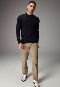 Polo negro de manga larga con tres botones, combinado con pantalones beige y zapatillas blancas con rayas negras. Textura suave, diseño ajustado.