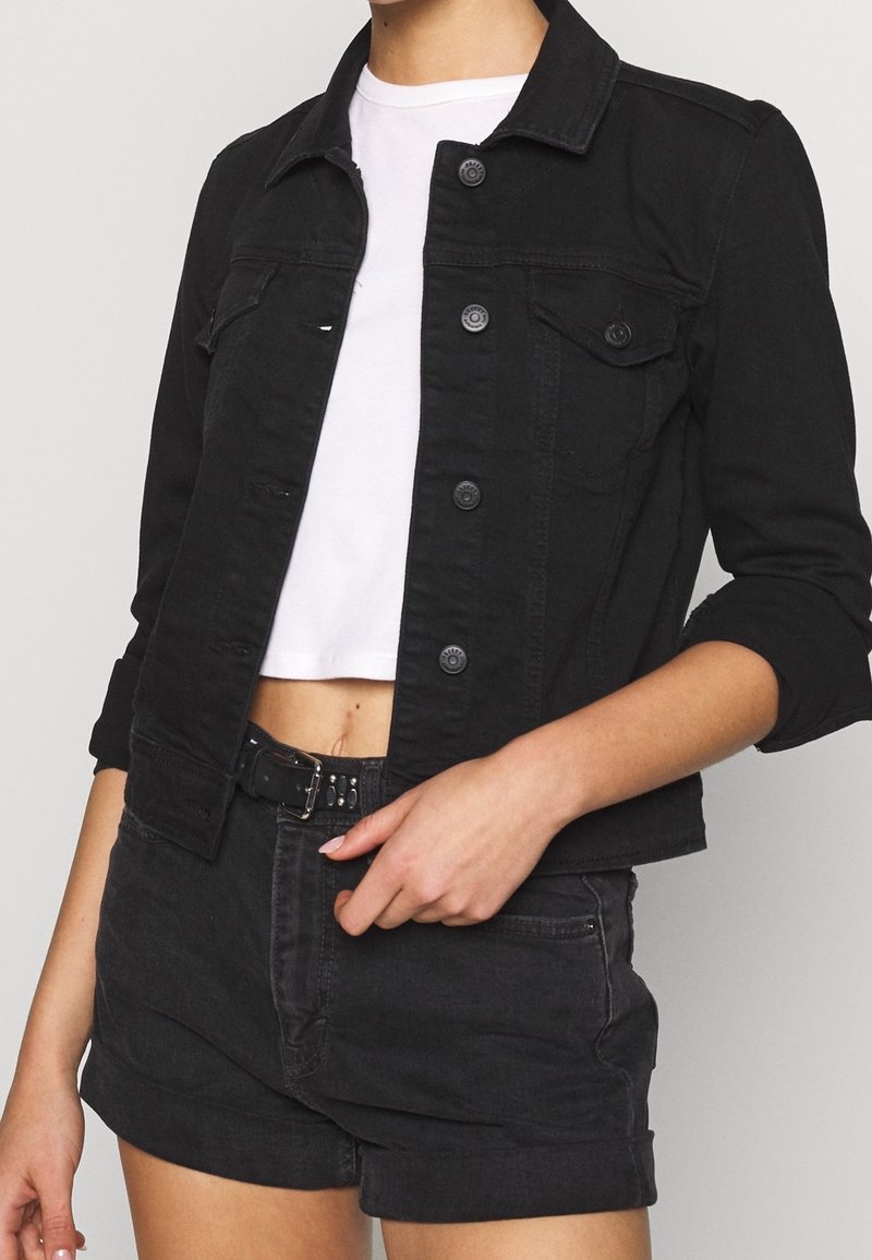 Femme portant une veste en jean noire à boutons par-dessus un t-shirt court blanc, assortie à un short en jean noir taille haute et une ceinture.