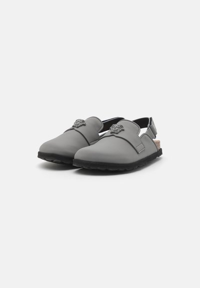 Hallid hallid kummist sussid slingback kujundusega, mille esiosas on lõvi embleem ja must tekstuuriga välistald haardeks.