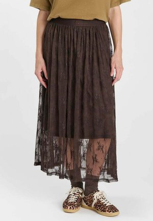 Persoon draagt een bruine kanten midirok, sneakers met luipaardprint, bruine sokken en een beige top, staande tegen een effen achtergrond.