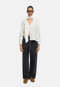 Biała bluzka w kropki z przewiązanym przodem, zestawiona z czarnymi spodniami typu wide-leg. Brązowe obuwie dopełnia całość. Minimalistyczny design i tekstura.