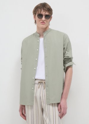 Camicia verde chiaro con bottoni e collo alla coreana. Presenta una texture liscia, indossata sopra una T-shirt bianca e pantaloni beige a righe.