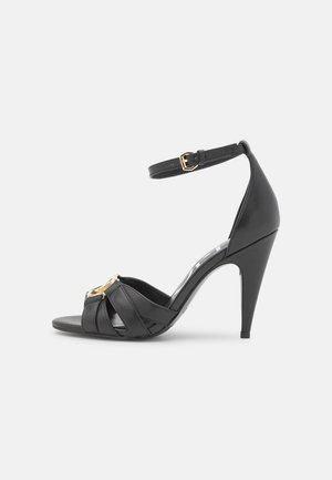 Schwarze High-Heels Sandale mit gekreuzten Riemen, verstellbarem Knöchelriemen und goldenen Hardware-Akzenten. Glattes Lederfinish, spitz zulaufendes Design.