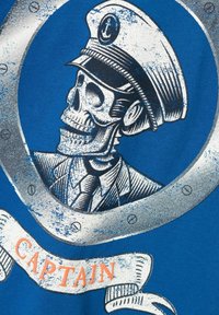 Un tejido azul presenta un gráfico de un esqueleto con un gorro de capitán y un diseño de portillo plateado. La pancarta debajo dice "CAPITÁN" en naranja.